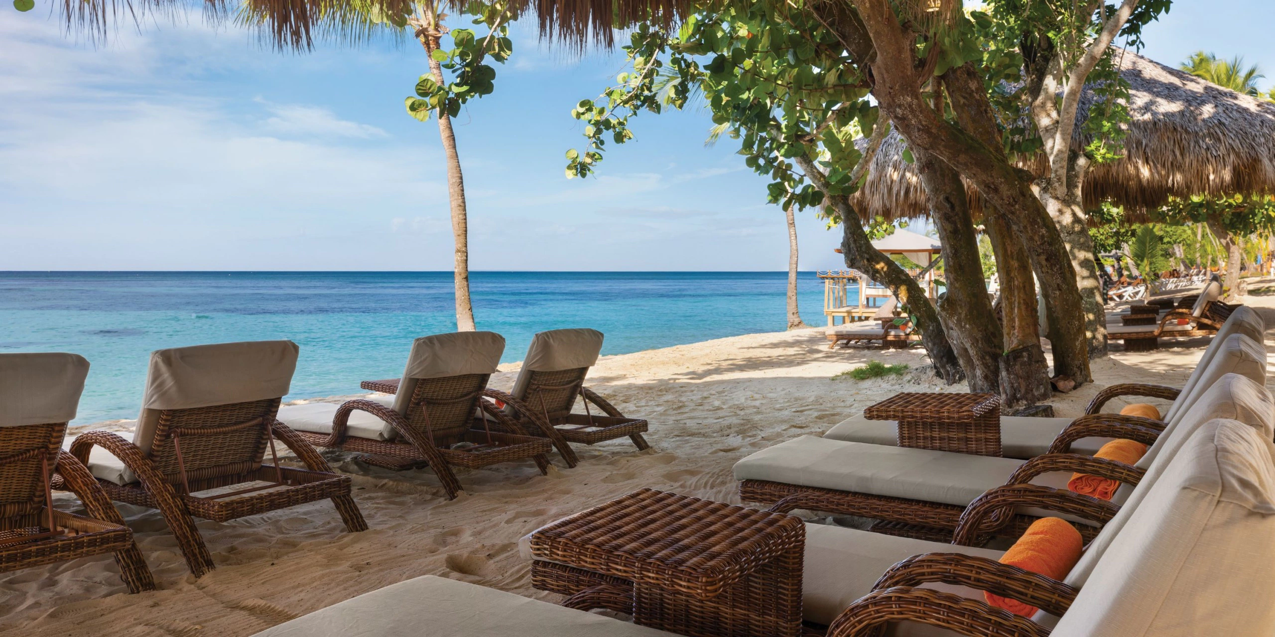 Beachfront lounge chairs beneath tropical shade trees at Secrets La Romana Resort & Spa.